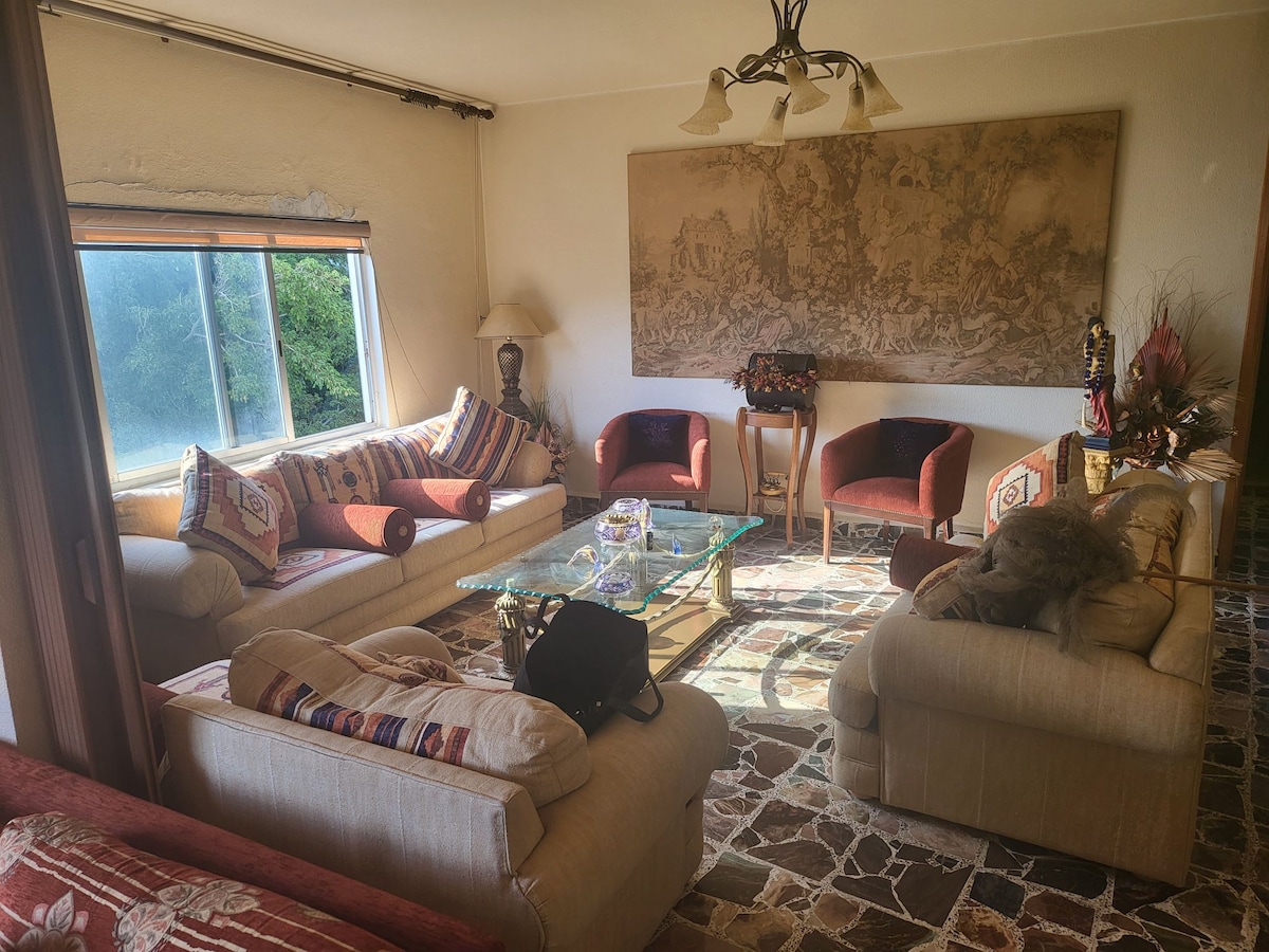 A cozy living area features comfortable sofas adorned with decorative cushions, complemented by two accent chairs. A glass coffee table sits at the center, with soft natural light filtering through large windows, highlighting the textured wall art above.