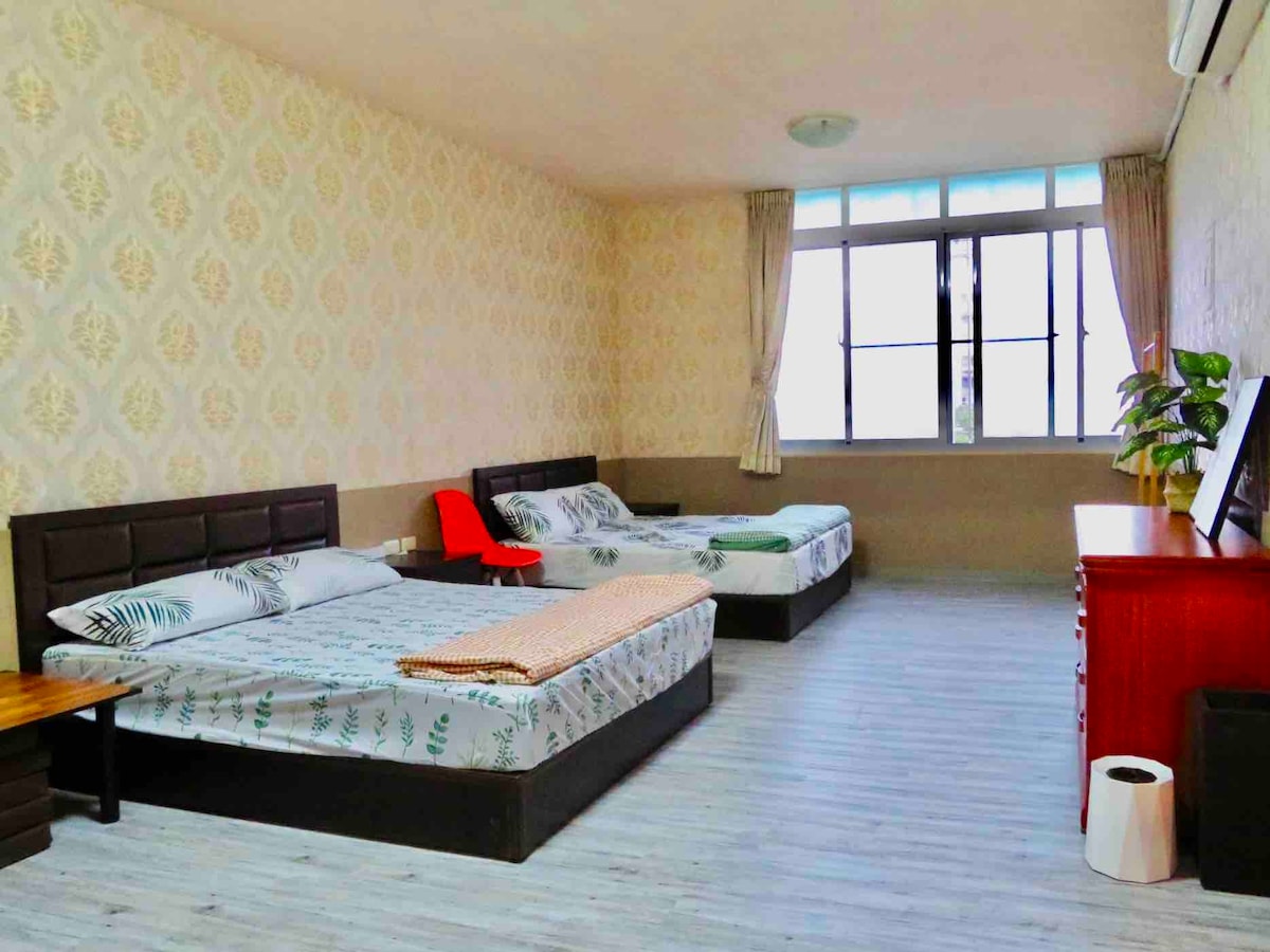 A spacious room features two large beds adorned with leaf-patterned bedding. Natural light streams in through a large window, while a red chair and a wooden table enhance the functional layout. A simple dresser and a small waste bin complete the welcoming atmosphere.