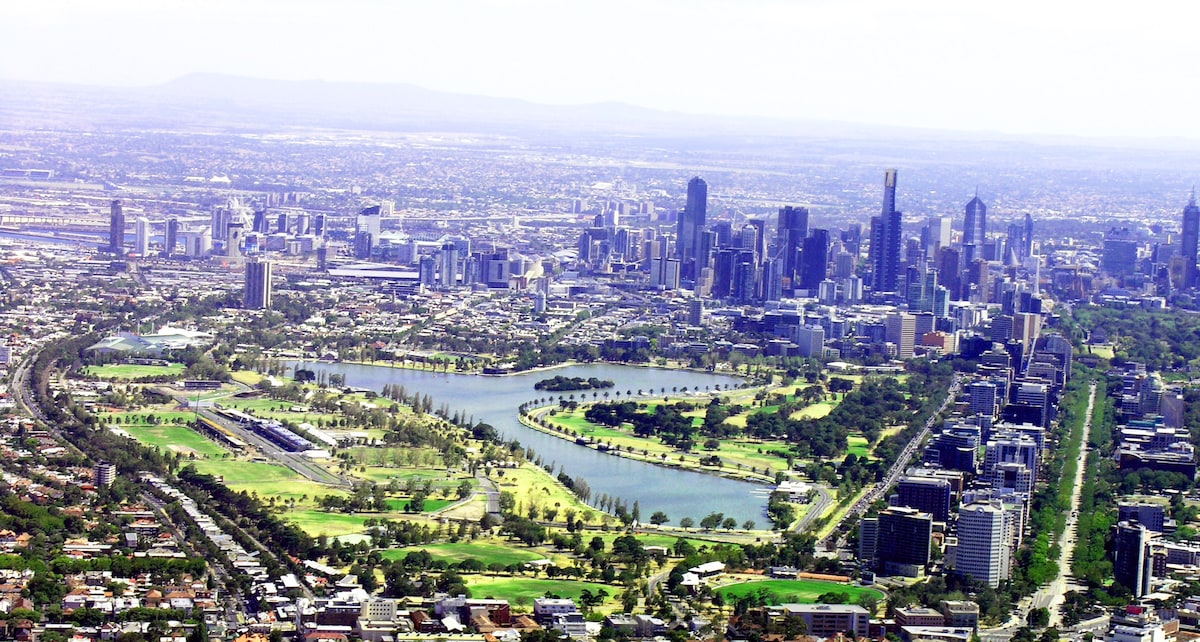 Aerial view captures a sprawling urban landscape with a mix of green spaces and modern architectural structures. A river meanders through the parkland, surrounded by lush trees, while the skyline of a vibrant city rises in the background against a clear sky.
