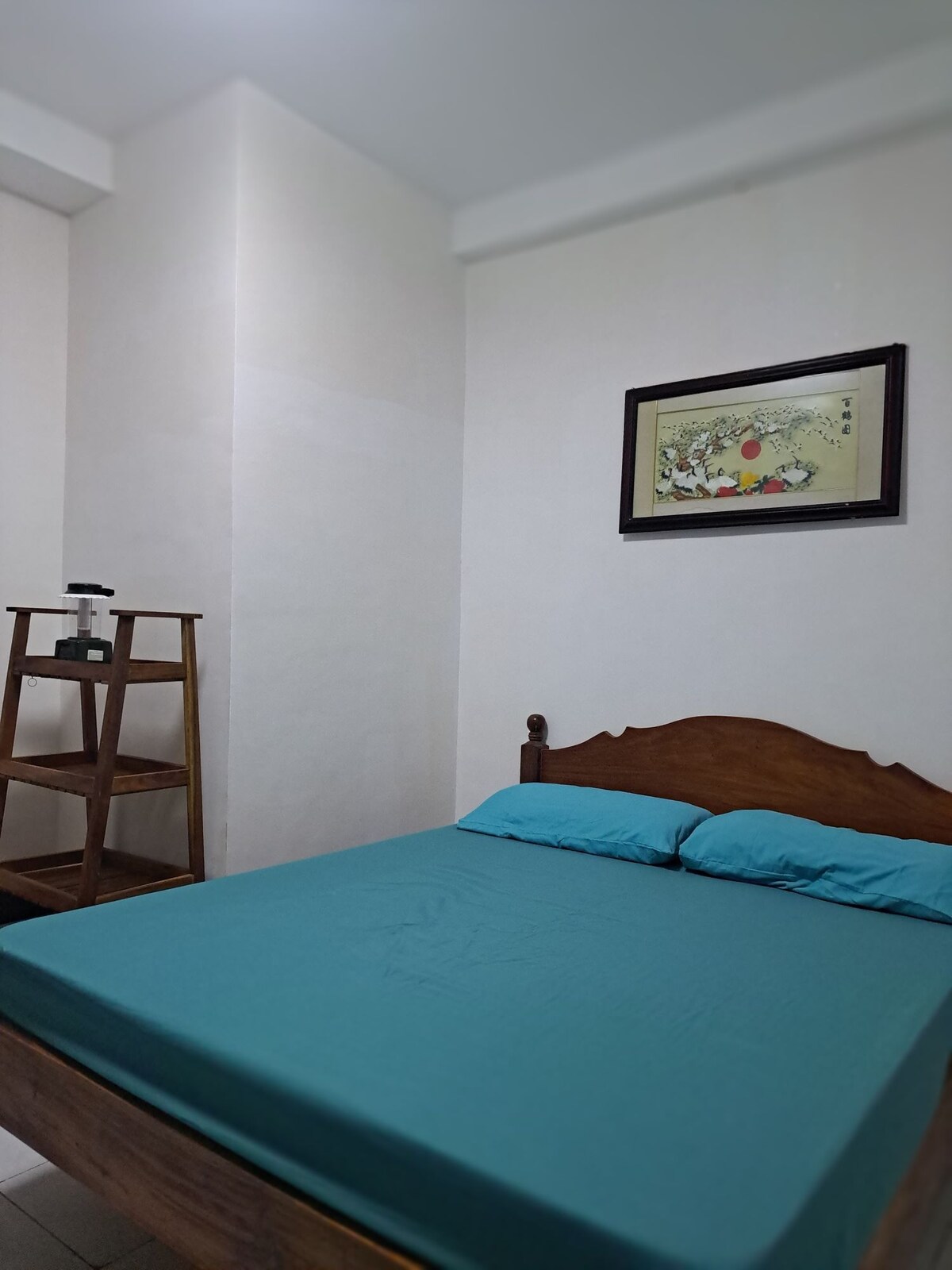 A cozy bedroom features a wooden bed with a teal bedspread and two matching pillows. A framed artwork adorns the wall above the bed. A wooden bookshelf is placed in the corner, adding functional charm to the space.