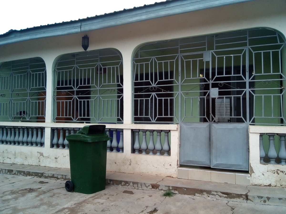 The exterior of the building features arched windows and doorways adorned with decorative metal grilles. A green garbage bin is positioned near the entrance, while the walls are painted in a soft green hue. A concrete pathway leads to the front door, which hosts a metal screen.