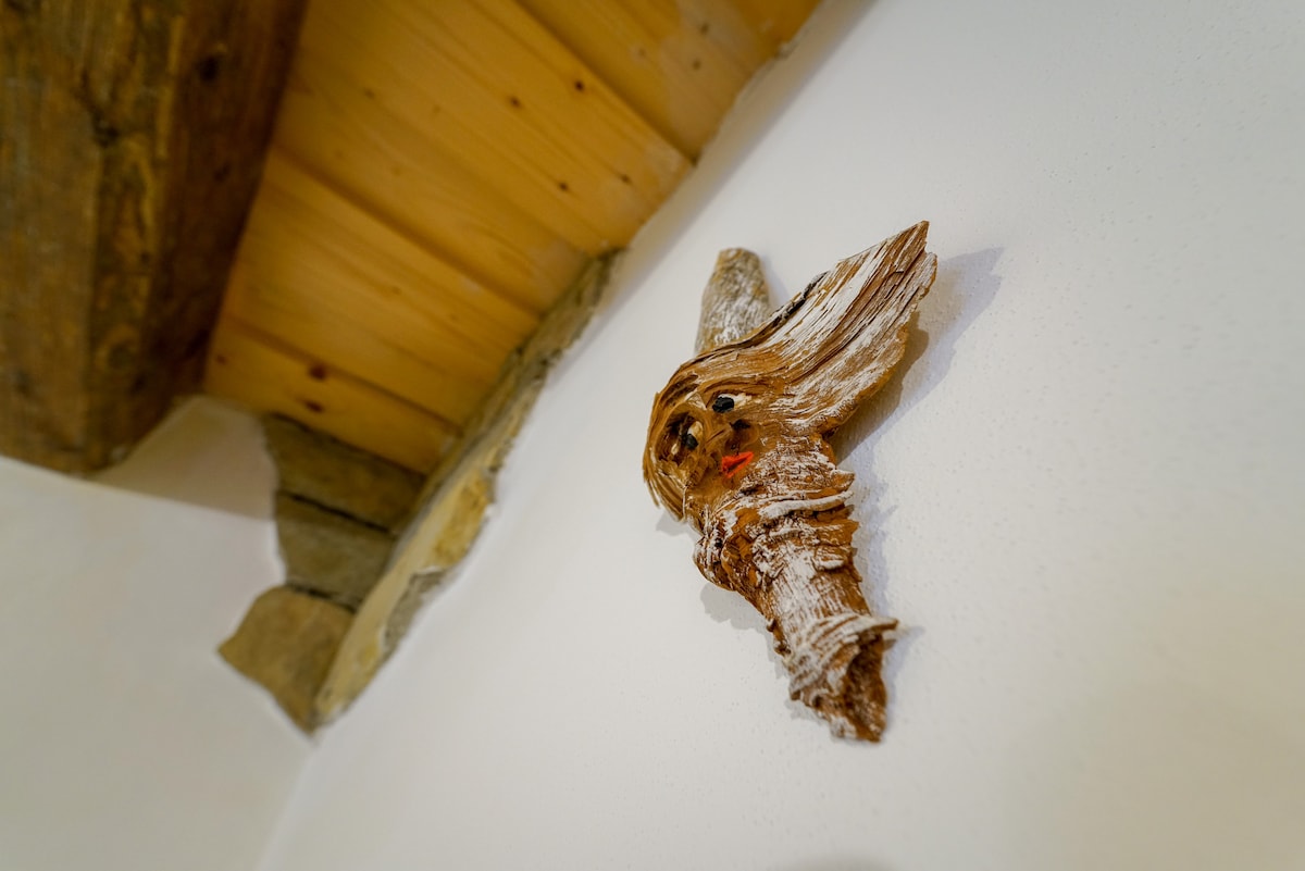 A decorative wall piece, crafted from natural materials, is mounted on a white wall. The piece features intricate textures and a blend of earthy tones, adding a unique touch to the interior. Wooden beams and stone elements can be seen in the background.