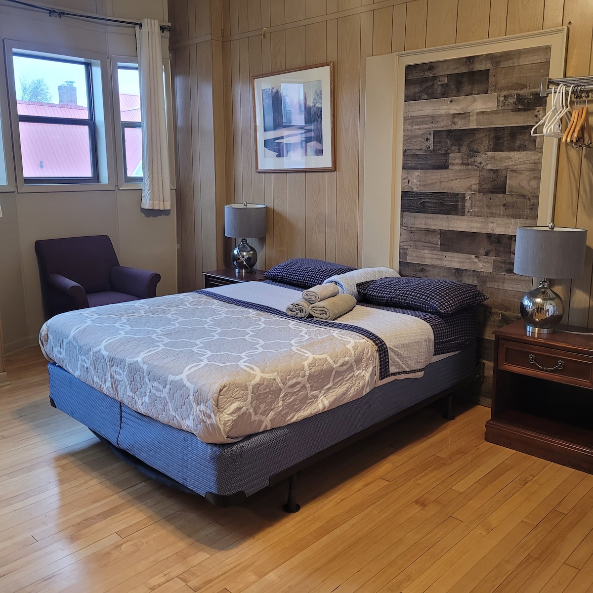 A well-appointed bedroom features a spacious bed with neatly arranged towels. A wooden accent wall enhances the design. A comfortable armchair sits in the corner, accompanied by two bedside tables with lamps. Natural light spills in through a nearby window.