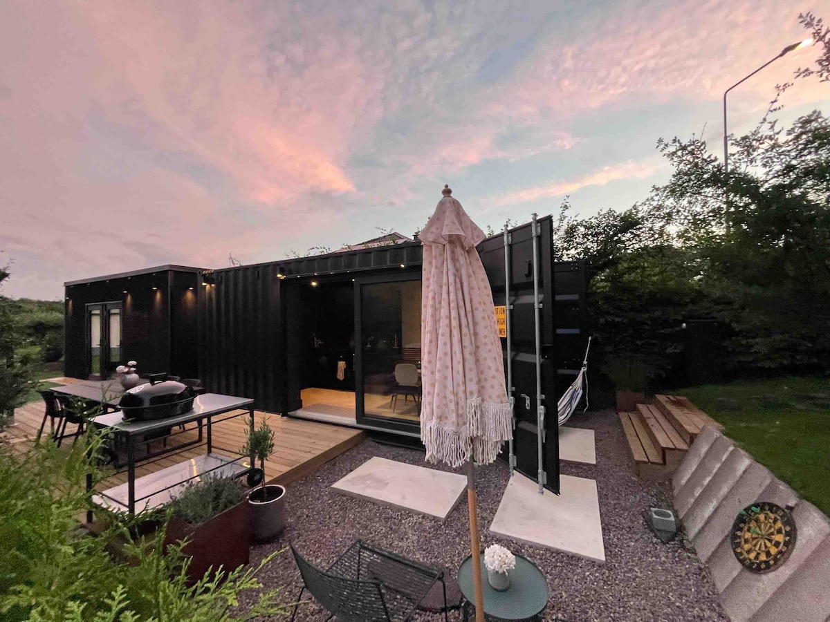 A container house exterior is showcased during sunset, featuring large glass doors that create a seamless connection with the surroundings. Outdoor furniture, including a dining table and chairs, are arranged on a gravel patio. A large umbrella provides shade, enhancing the outdoor living experience.