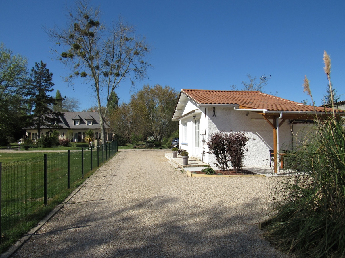 A gravel pathway leads to the charming white house with a terracotta roof. Surrounding greenery includes trees and shrubs, creating a tranquil setting. A neighboring building can be seen to the left, contributing to the serene atmosphere of the property.