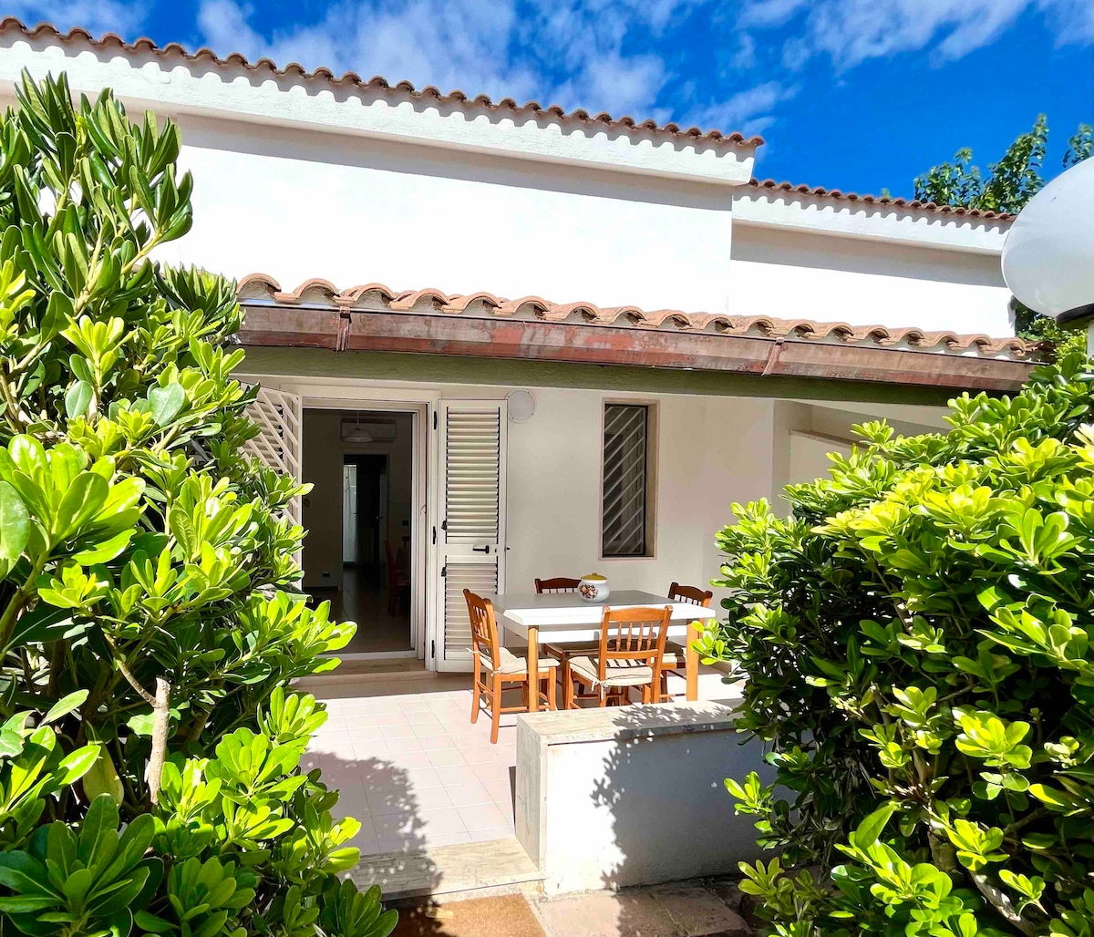 An outdoor patio area features a wooden dining table surrounded by chairs, nestled among lush greenery. The entryway to the home is visible, revealing a glimpse of the interior. A bright blue sky is visible above, enhancing the serene setting.