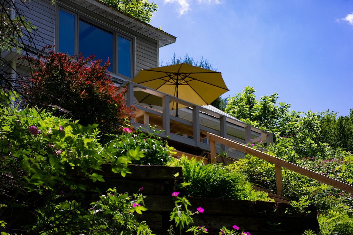 A new scenic deck is visible from below, featuring a large yellow umbrella providing shade over a wooden table. Surrounding greenery includes vibrant flowers and lush foliage, creating a serene outdoor space. A charming view of the house is complemented by a clear blue sky.