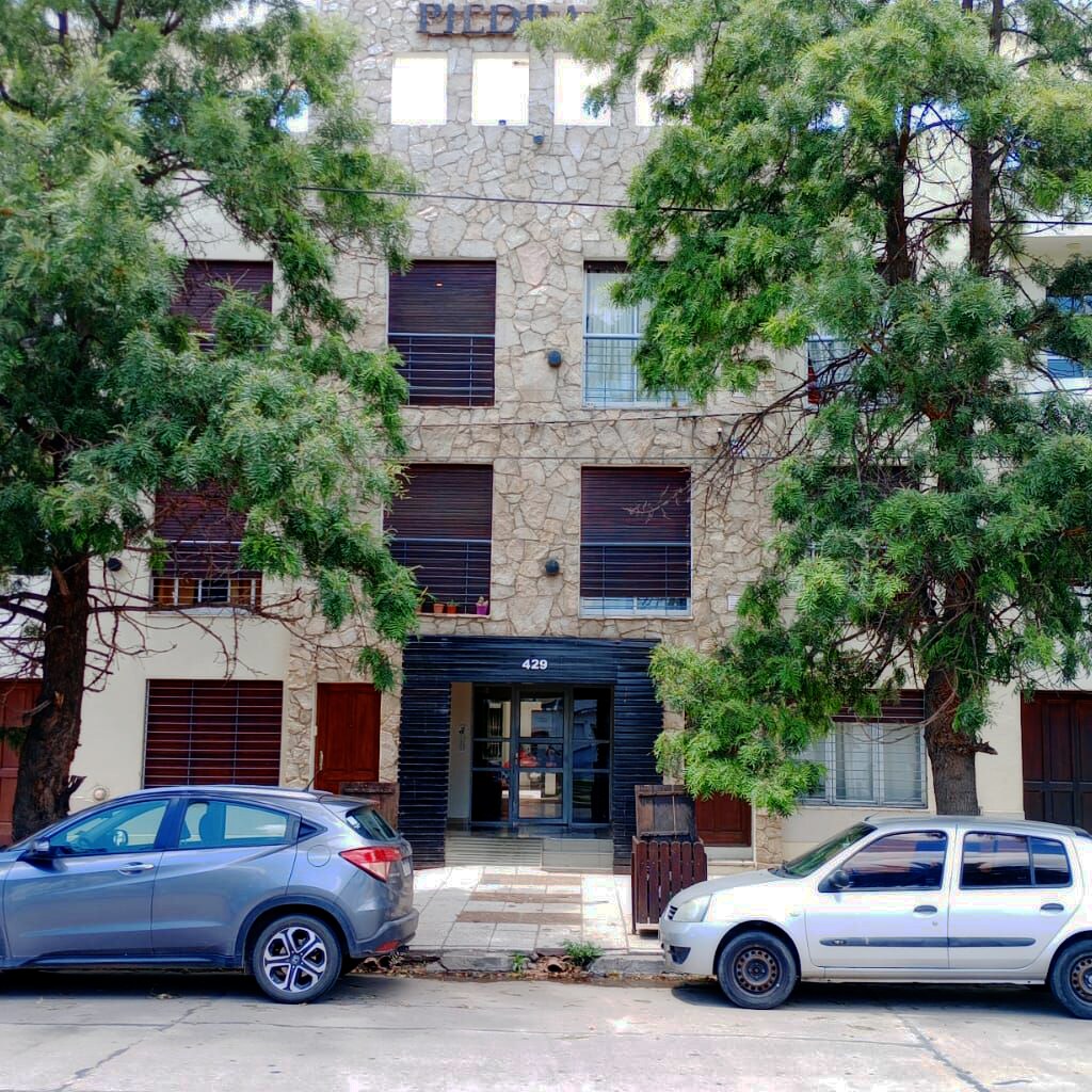 The building showcases a stone exterior with multiple floors, featuring dark shuttered windows and a central entrance. Surrounding trees provide shade, while parked cars are visible along the street, contributing to the urban atmosphere.