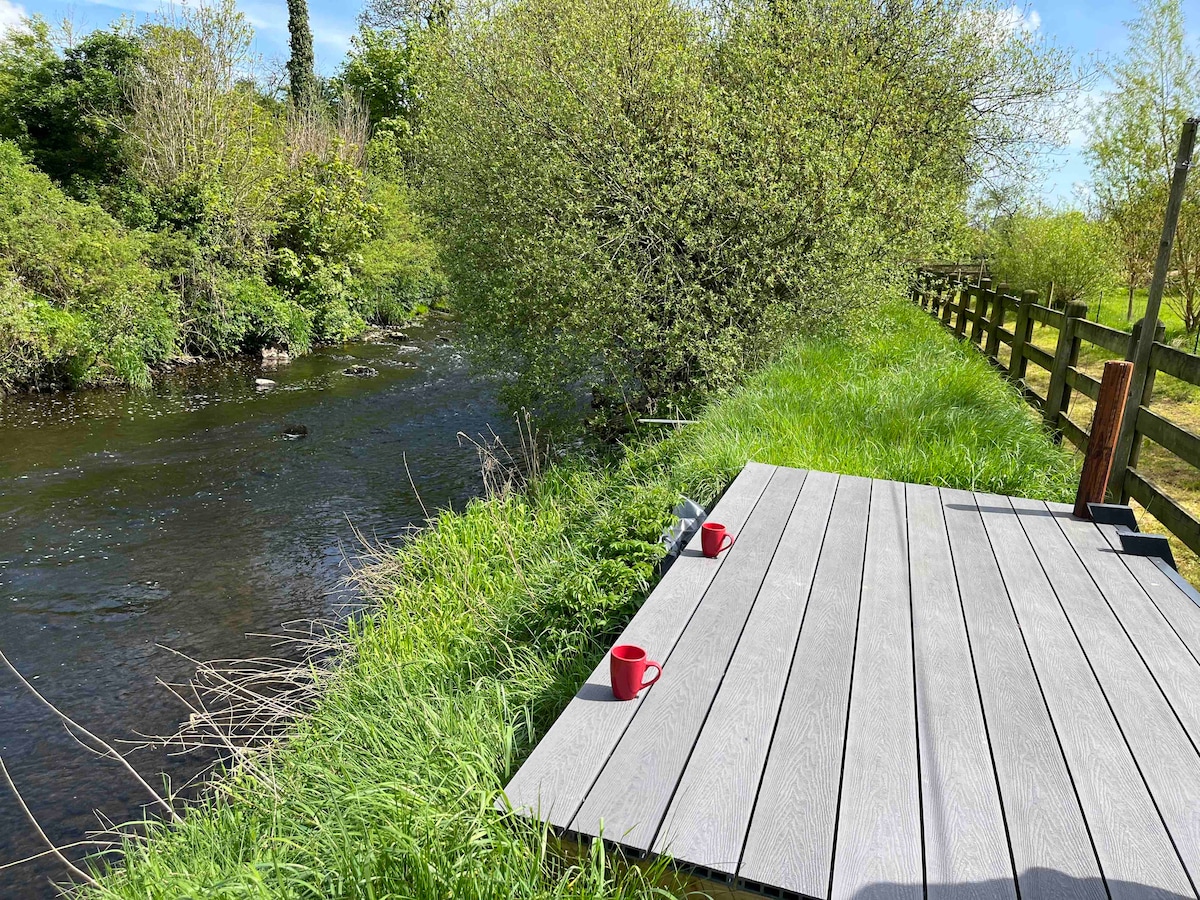 A wooden deck is set along the banks of a flowing river, surrounded by lush greenery. Two red cups are placed on the deck, enhancing the tranquil setting. A wooden fence lines the area, complementing the natural landscape.