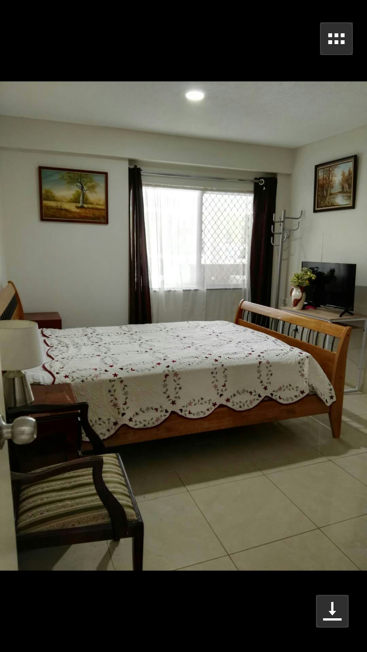 A spacious master room features a queen bed with a decorative blanket. Natural light enters through a large window adorned with dark curtains. A wooden dresser and a chair are positioned beside the bed, with wall art complementing the decor. An electric television is mounted on the wall.