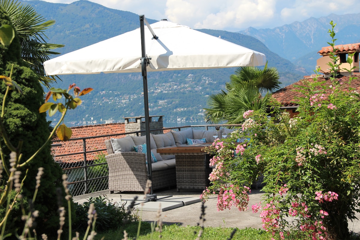 A shaded lounge area is situated on a terrace, featuring comfortable seating and a large umbrella. Lush greenery and colorful flowers surround the space, with distant mountains visible in the background, creating a serene outdoor setting.