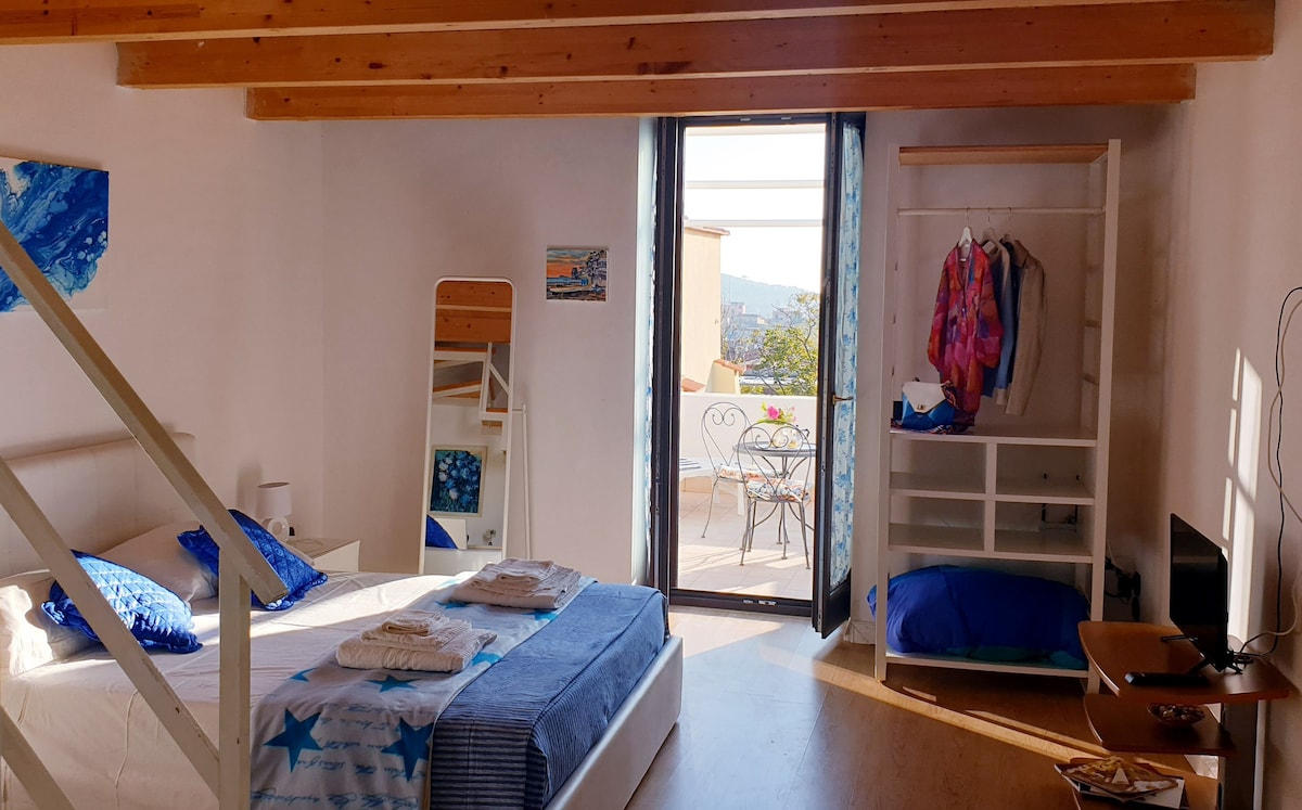 A bright bedroom features a comfortable bed covered with soft linens. Natural light fills the room through a sliding glass door that opens to a terrace. The space includes a mirror, open shelving for clothing, and a TV, making it well-equipped for guests.