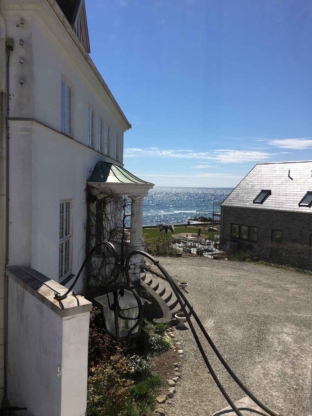 The image captures a sunny view of the sea, seen from a property featuring a white facade and a green roof. Spiral stairs lead down to a gravel area adorned with plants, while a pathway directs towards the beach in the distance.