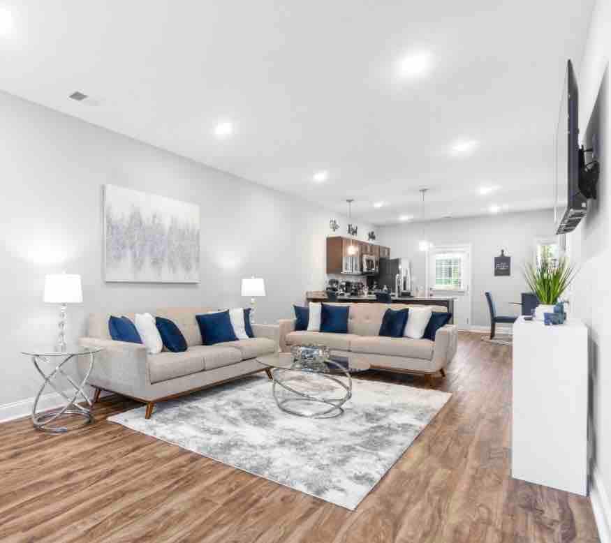 A modern living area showcases a light grey color palette with elegant furnishings. Two sofas are arranged around a circular glass coffee table on a patterned area rug. Soft lighting from table lamps enhances the inviting ambiance, while a kitchen area is visible in the background.