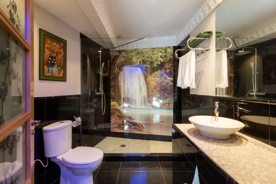 A modern bathroom features a sleek design with dark tiled walls and floor. A large, round basin is positioned on a textured countertop, while a shower area includes a waterfall mural as a backdrop. Essentials like towels are neatly arranged for convenience.