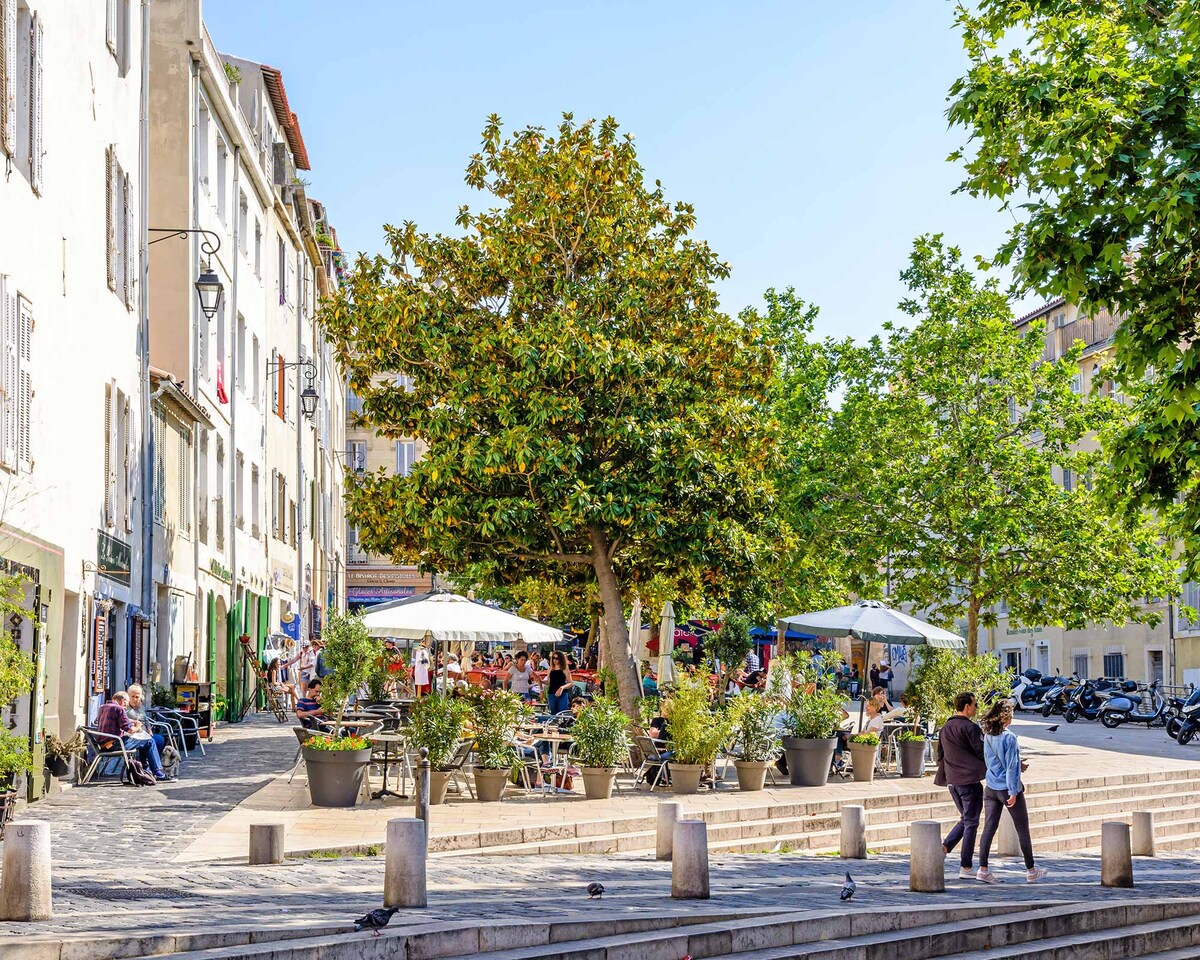 A lively square is surrounded by charming buildings, featuring large trees and potted plants. Outdoor seating under umbrellas accommodates visitors. Groups of people can be seen enjoying their time, with a mix of greenery and urban elements creating a welcoming atmosphere.