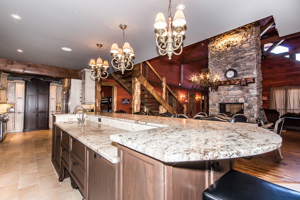 A spacious kitchen featuring a large granite island with seating. Elegant light fixtures hang overhead, illuminating the area. The backdrop showcases a rustic stone fireplace and a wooden staircase that leads to a second level, enhancing the inviting ambiance of this open-concept space.