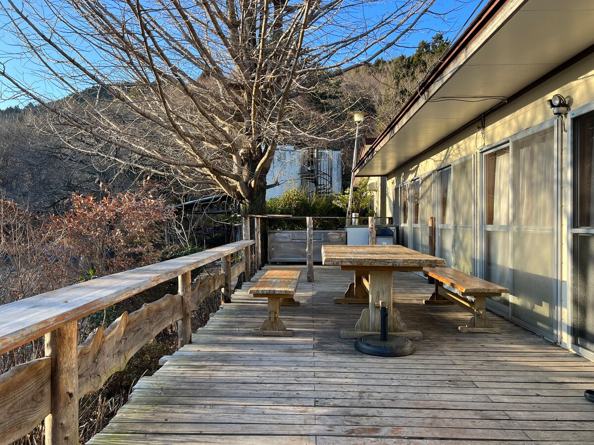 A spacious wooden deck is seen, furnished with picnic tables and benches. A large tree is situated nearby, its branches bare. The setting indicates a serene outdoor area, surrounded by nature and offering a peaceful spot for relaxation.