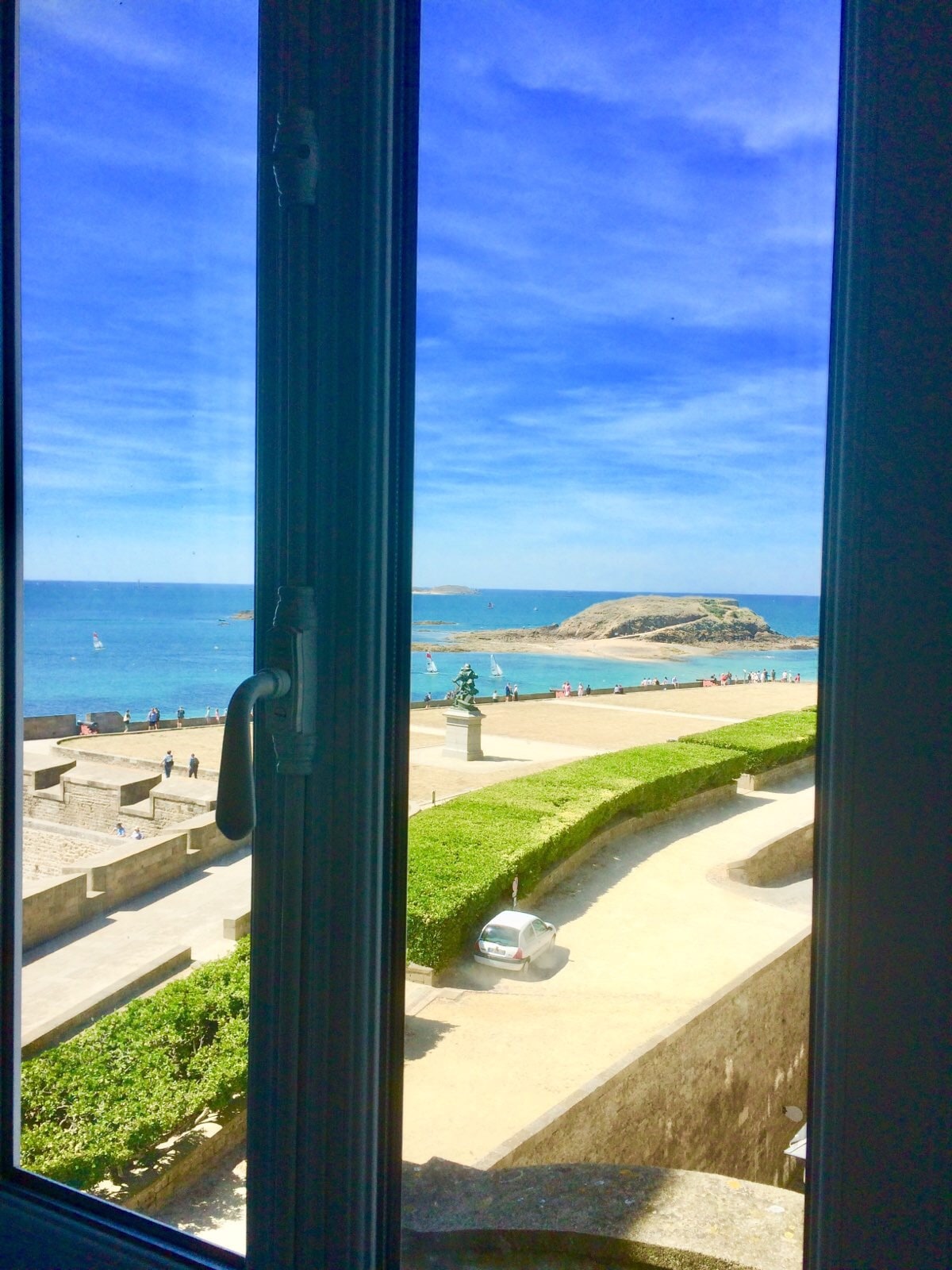A scenic view is captured through a window, showcasing the blue sea and a distant island. The foreground features a green hedge along a coastal pathway, inviting passersby to enjoy the tranquil surroundings.