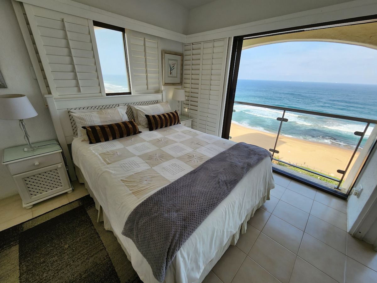 A well-appointed bedroom features a spacious bed with a decorative quilt and neatly arranged pillows. Large windows open to stunning ocean views, while light streams in through the shutters. A bedside lamp and a small nightstand complement the coastal ambiance of the room.