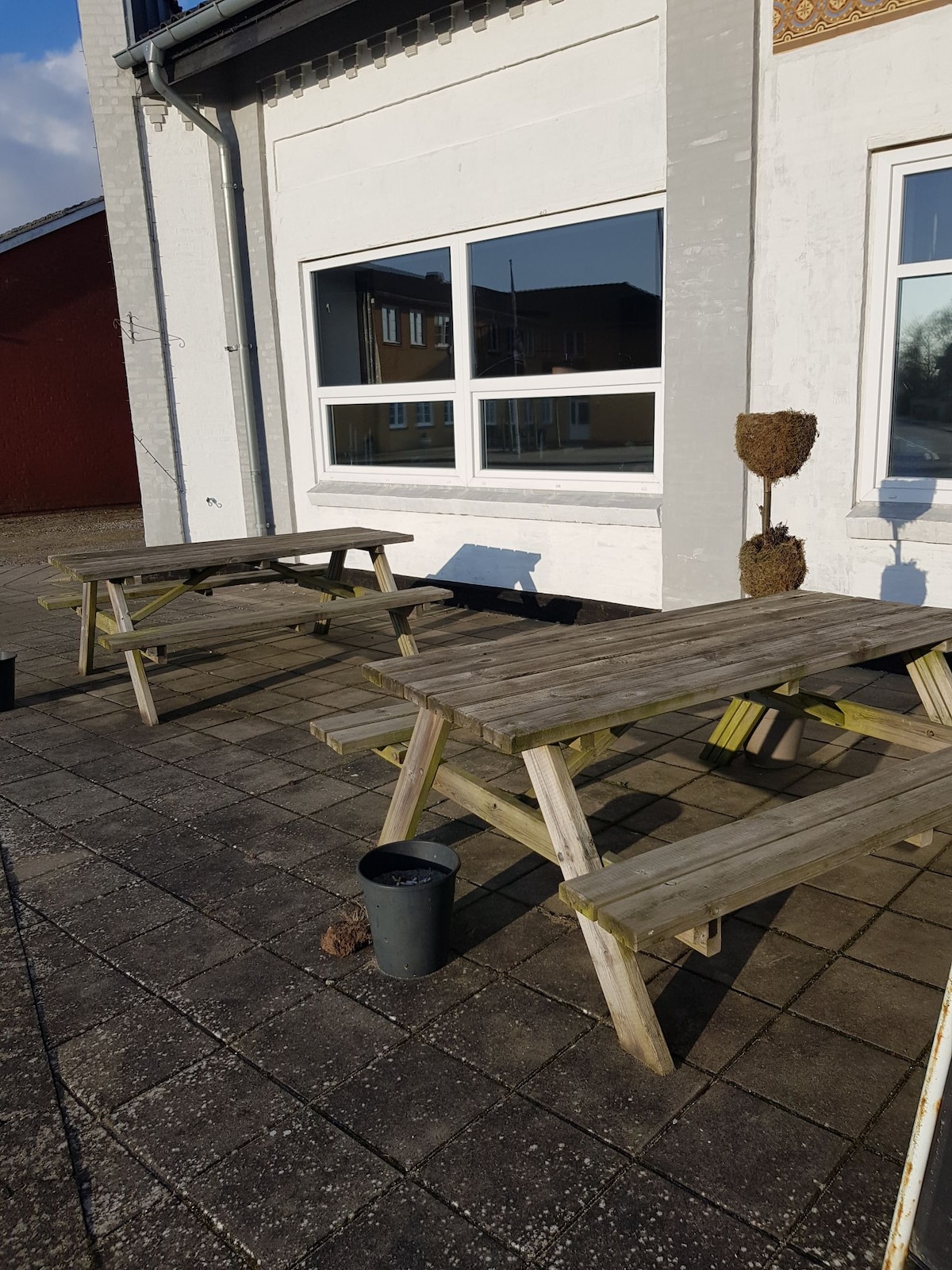 An outdoor area features two wooden picnic-style tables positioned on a paved surface. A large window is visible in the background, allowing natural light to illuminate the space. A plant pot is placed near one of the tables, and natural surroundings can be seen beyond.