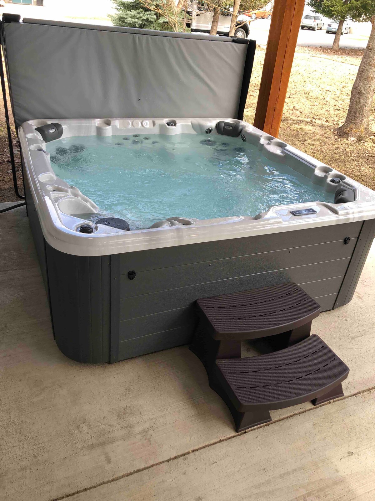 A hot tub filled with clear water is situated on a concrete surface, featuring steps for easy access. The surroundings include natural shade from nearby trees, enhancing the inviting outdoor space.