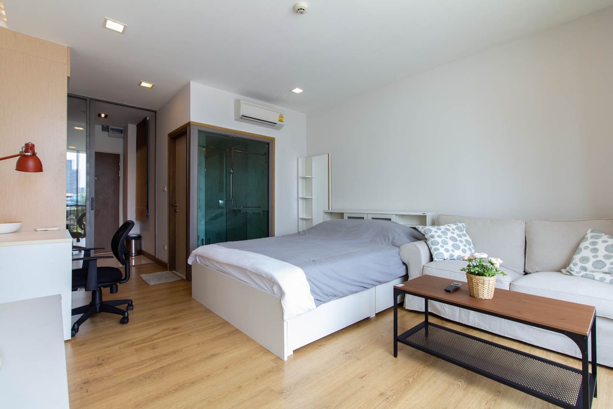 A spacious studio layout features a comfortable bed with neutral bedding, accompanied by a light-colored sofa. A small table sits nearby, and a desk with a chair is positioned against the wall. Natural light flows through the glass door leading to a balcony.