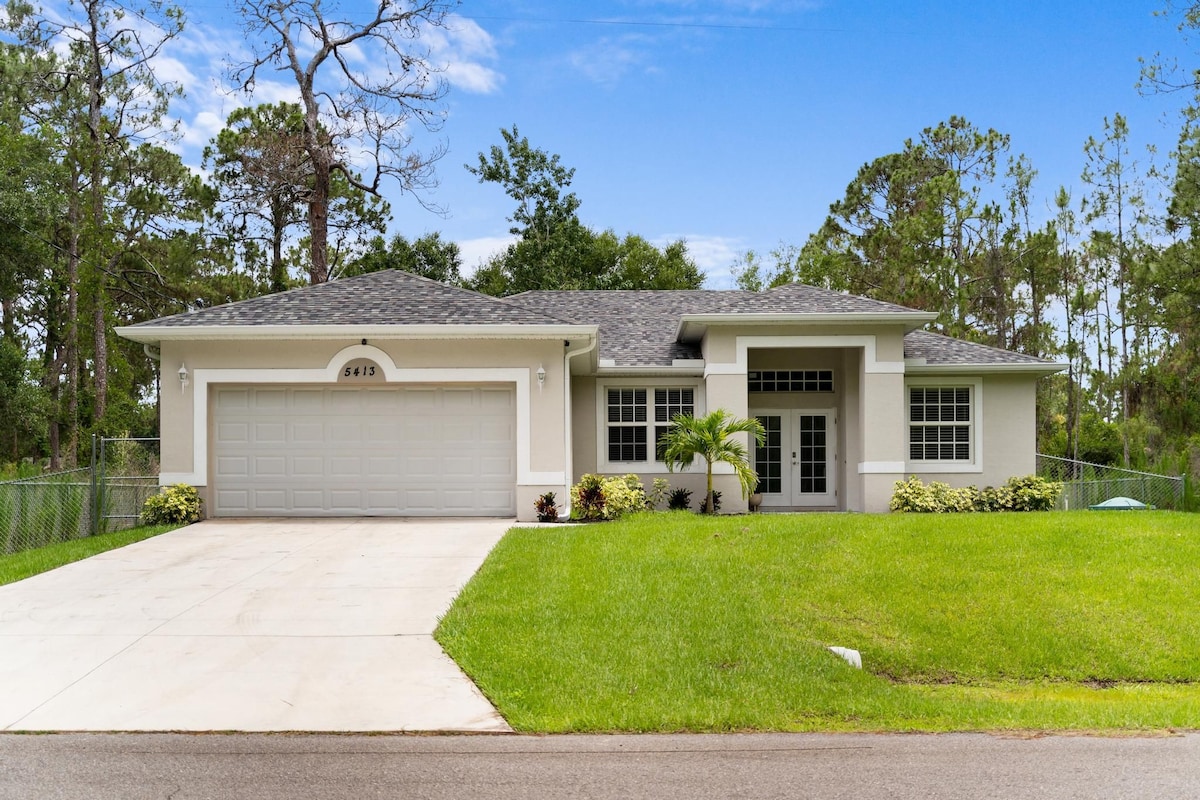 A modern single-story home is showcased with a spacious driveway and a well-maintained front yard. Lush greenery surrounds the property, and large windows illuminate the inviting exterior. The home features a covered entryway and a serene setting under a clear blue sky.