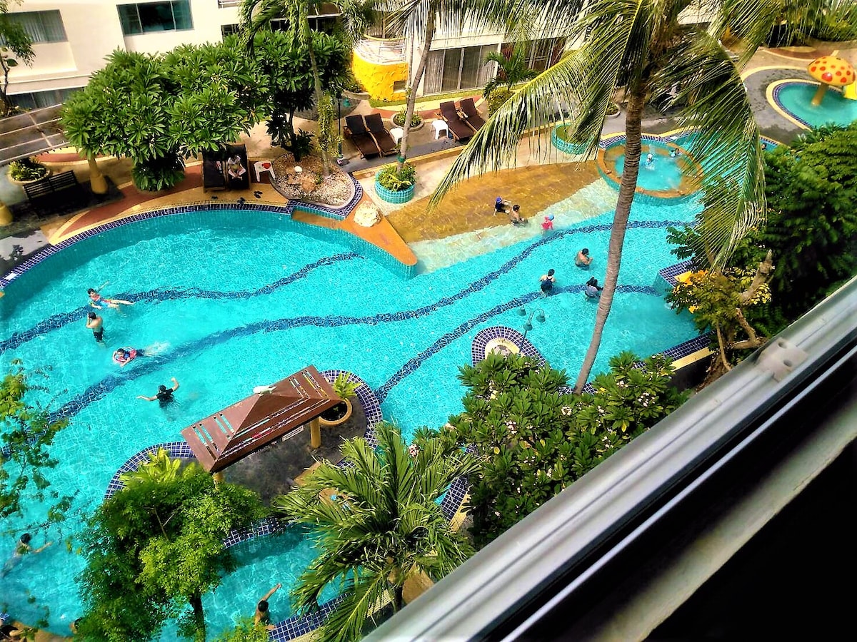 An aerial view of a resort-style pool area is displayed, featuring a winding blue pool surrounded by lush greenery and palm trees. Guests can be seen enjoying the water, while lounge chairs are arranged along the poolside, creating inviting spaces for relaxation.