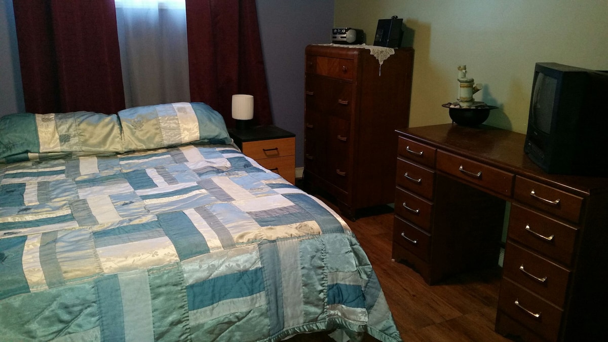 The cozy bedroom features a double bed with a light blue and white quilted cover. A dresser and a desk with several drawers provide ample storage. Natural light enters through a window, partially covered by burgundy curtains, adding a touch of warmth to the space.