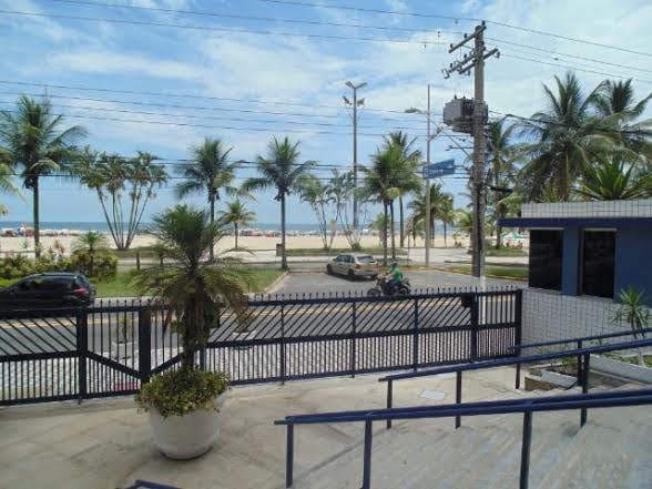 A view from the property shows a sandy beach across the street, framed by palm trees. The scene features a clear sky and gentle waves in the distance. A decorative planter is positioned at the entrance, adding greenery to the welcoming atmosphere.