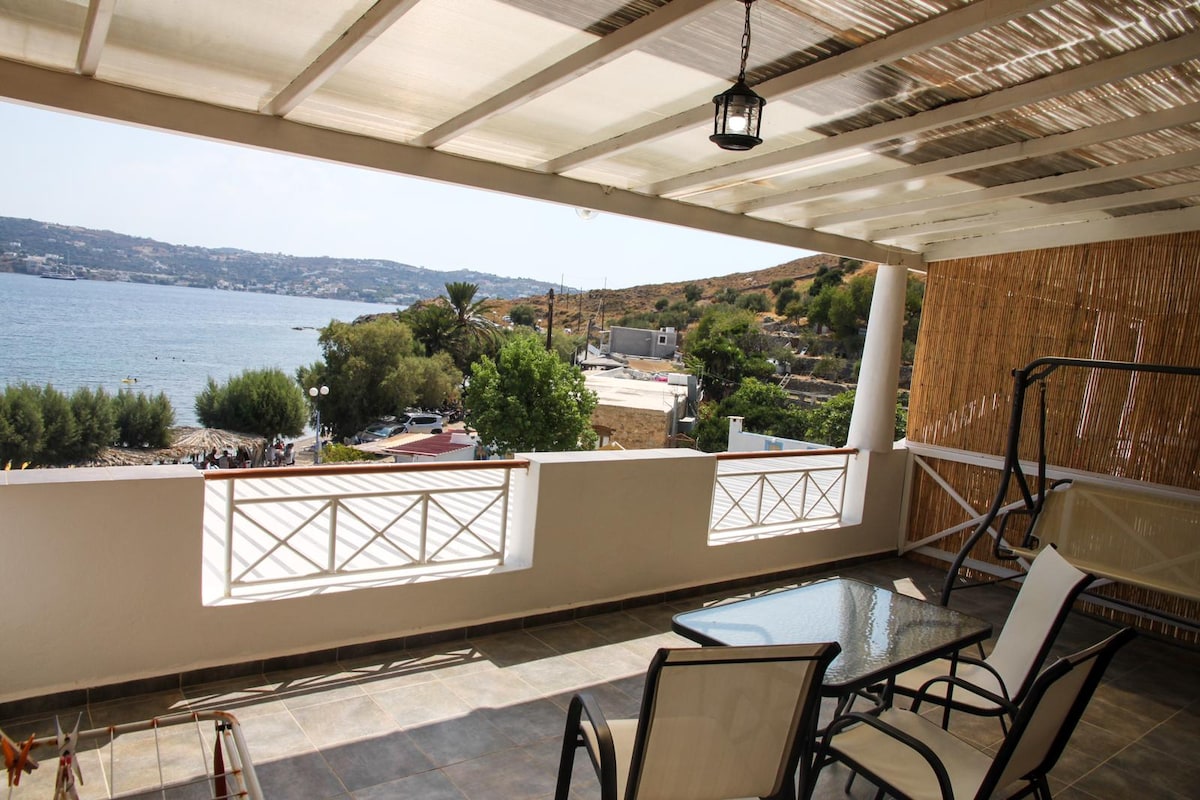 A covered terrace is visible, featuring two chairs and a small glass table. The serene view of the sea and hillside is emphasized by an open design, allowing natural light to fill the space. A soft canopy overhead adds to the ambiance.