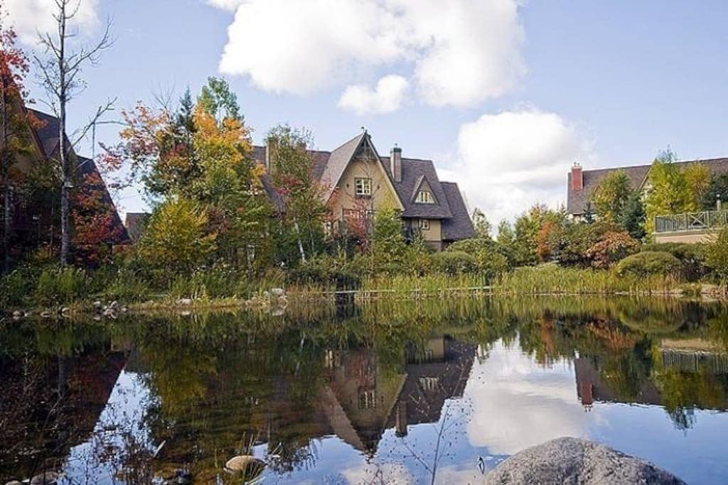 A tranquil landscape features a serene pond with reflections of charming multi-story buildings surrounded by vibrant autumn foliage. Lush greenery lines the water's edge, complementing the natural beauty of the scene under a partly cloudy sky.
