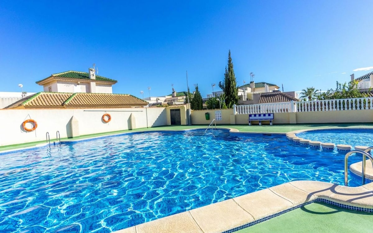 A large swimming pool is prominently featured, surrounded by bright blue water reflecting the clear sky. Spa-like seating areas are available near the water, with built-in steps leading into the pool. Lush greenery and surrounding buildings create a welcoming environment.