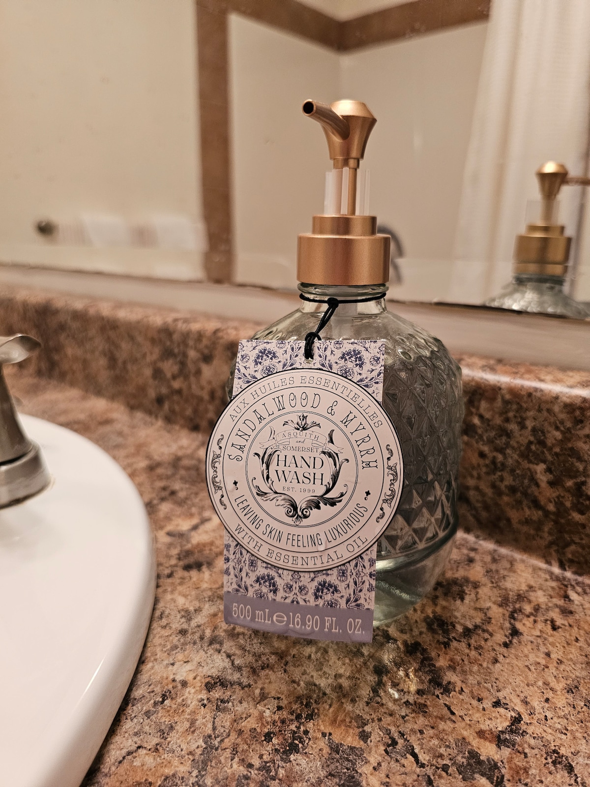 A decorative hand wash dispenser is positioned on a granite counter. The glass bottle, embossed with a diamond pattern, features a gold pump and a lavender label indicating a sandalwood and vanilla scent. A stylish sink is seen in the background.