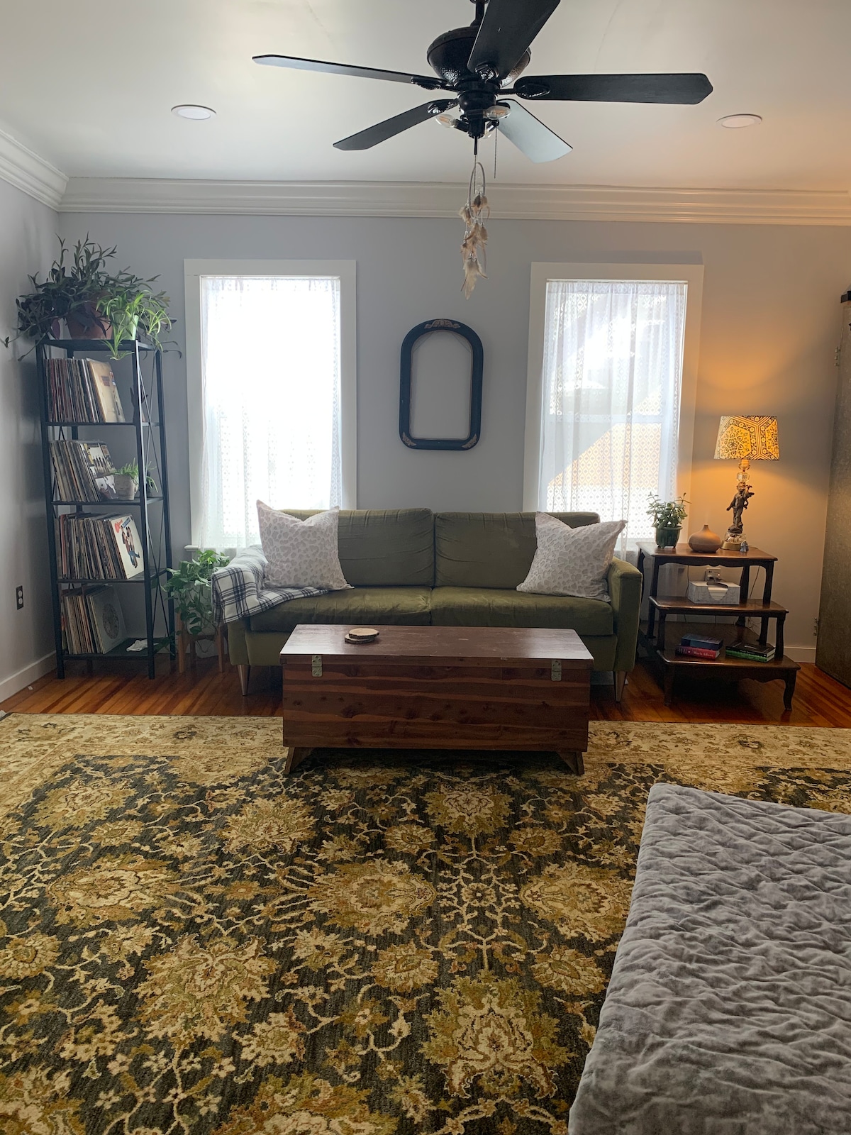 A cozy living area features a green sofa with decorative pillows, accompanied by a wooden coffee table. Natural light filters through two nearby windows, highlighting a warm area rug. A bookshelf filled with vinyl records and plants adds character to the space.