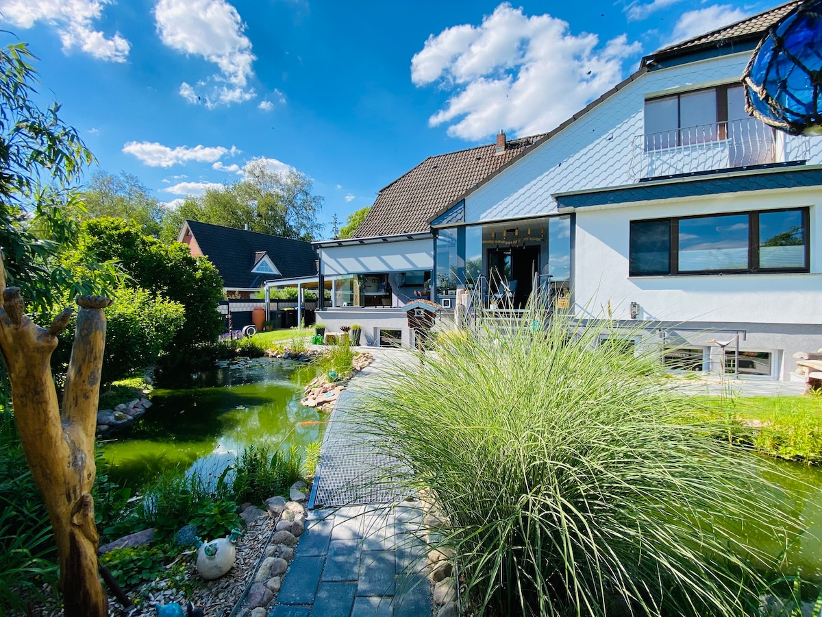 The exterior of the holiday apartment is depicted, showcasing a tranquil garden featuring a pond surrounded by lush greenery. A stone pathway leads to the entrance. The building is partially covered by a canopy, with large windows allowing natural light into the interior.