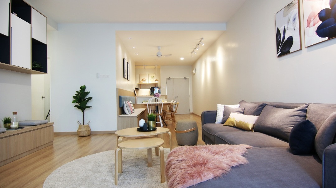 A bright and spacious living area is depicted, featuring a comfortable gray sofa with decorative pillows and a soft pink throw. A round coffee table sits on a large area rug, while a dining table and kitchen are visible in the background. Wall art and a houseplant add warmth to the space.
