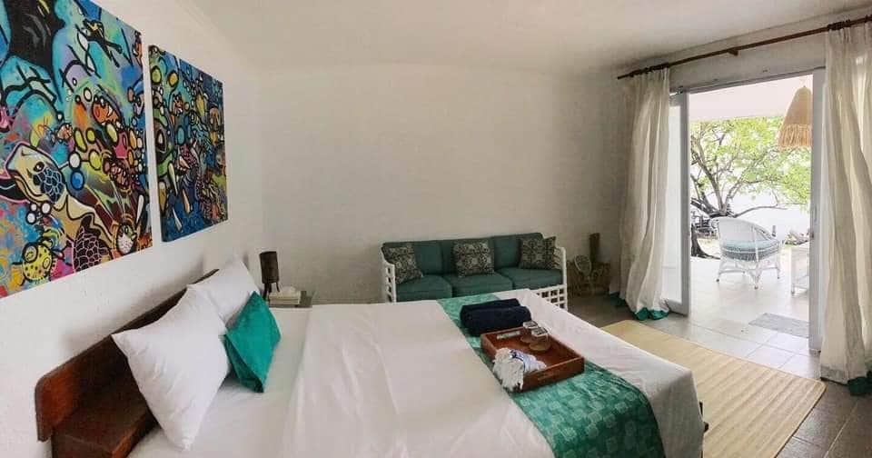 The bright sleeping area features a spacious bed with crisp linens and decorative pillows. Colorful artwork adorns the walls, while a comfortable seating area is positioned beside glass doors. Natural light floods the room, complemented by soft curtains and a soothing color palette.