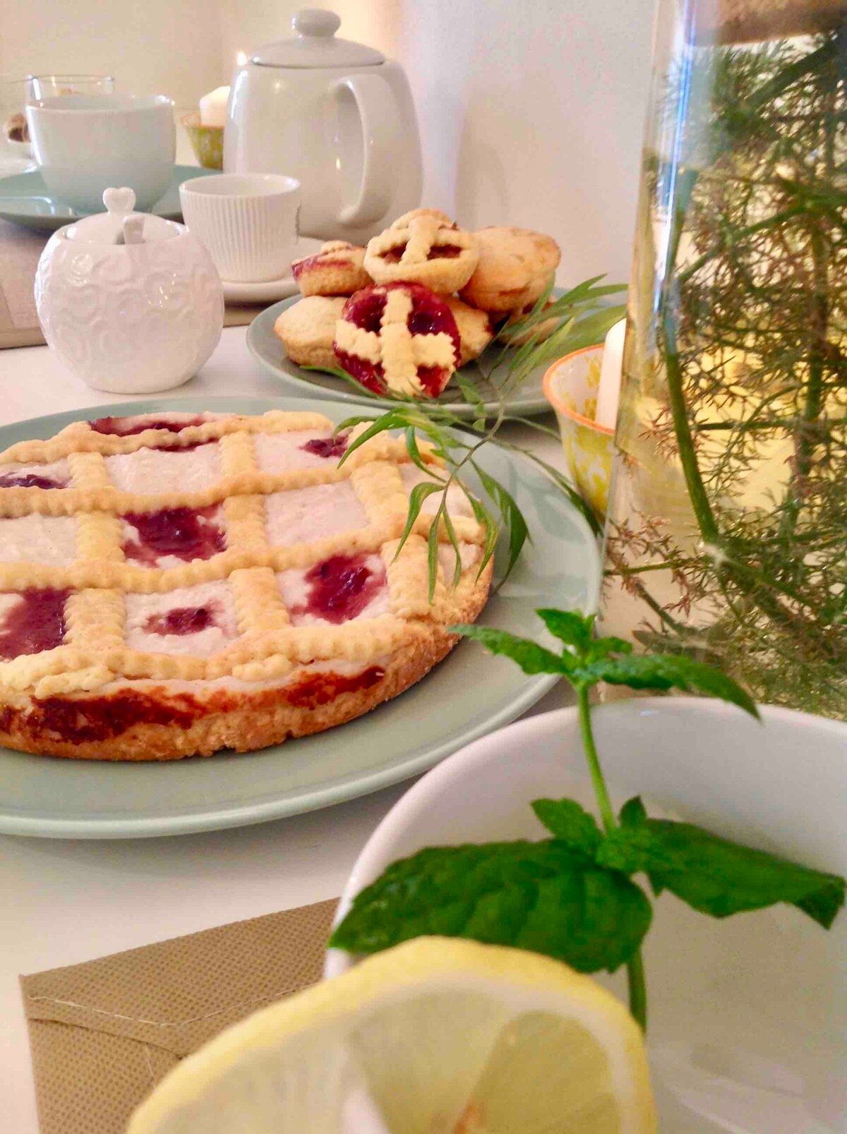 A table setting is presented with a variety of baked goods. A pie topped with a crisscross design and filled with fruit preserves sits alongside small cookies. A teapot and cups are arranged, accompanied by a glass of herbal tea infused with fresh mint and lemon.