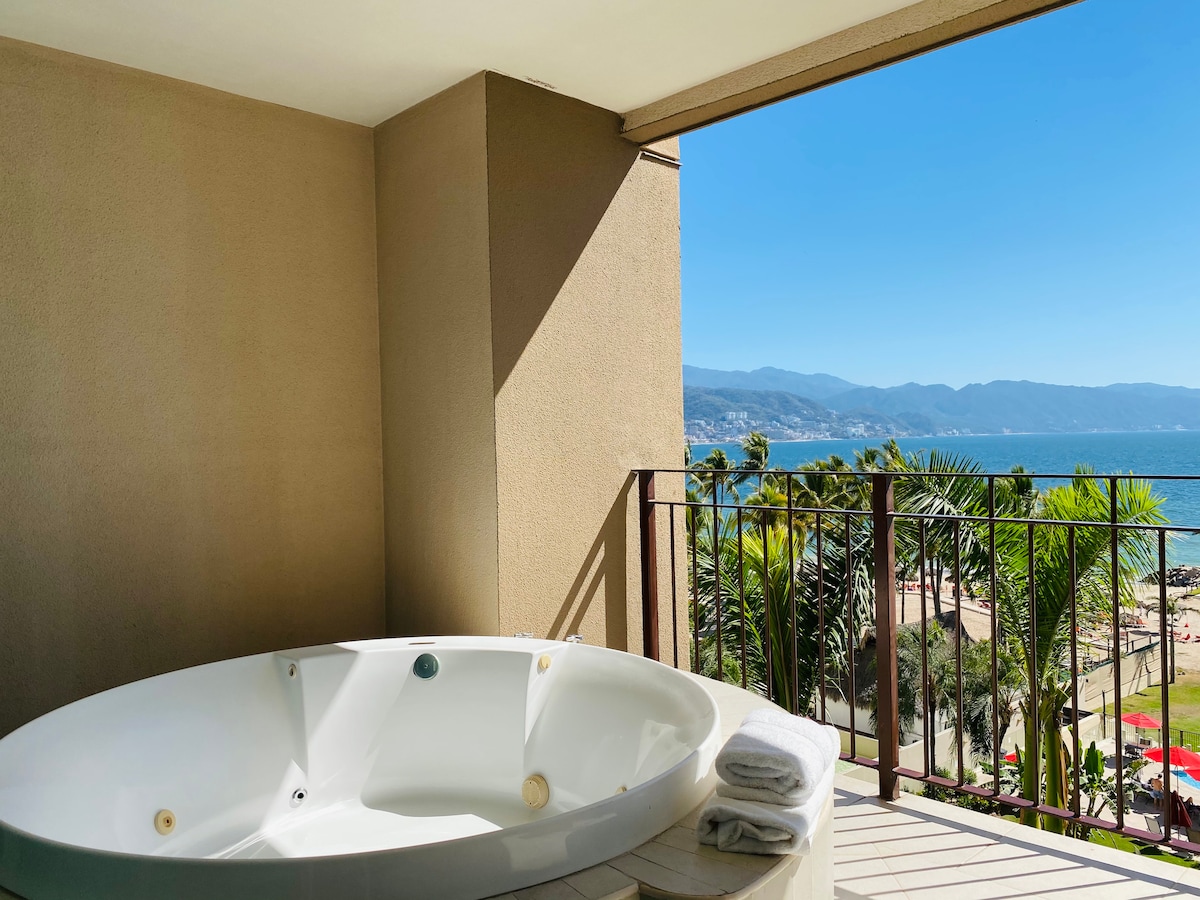 A private balcony features a round, white Jacuzzi overlooking the ocean. Green palm trees frame the view, while the blue water extends to the horizon, complemented by distant mountains. Soft white towels are neatly placed nearby.