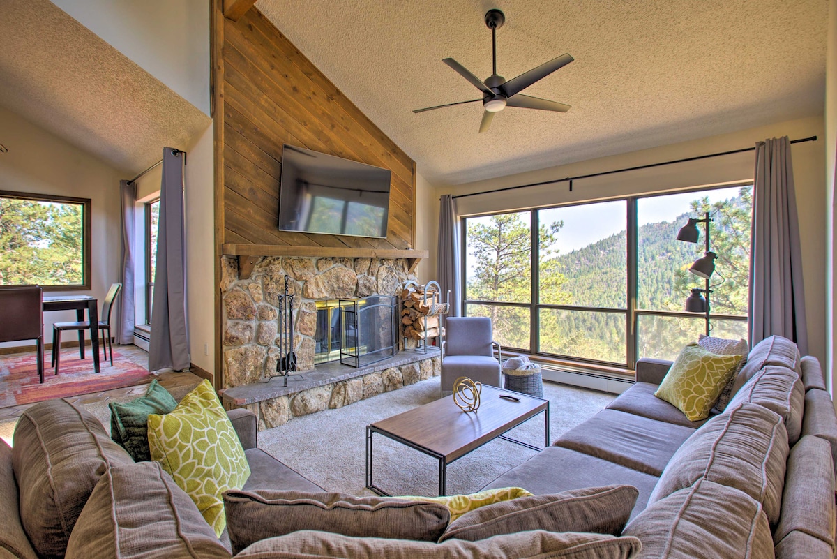 A spacious living room features a large L-shaped sofa adorned with decorative pillows. A stone fireplace is situated below a wall-mounted smart TV, and large windows showcase mountain views, allowing natural light to fill the space. A dining area is visible in the background.