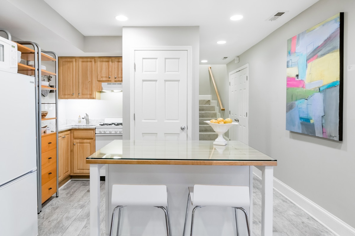 A modern kitchen and dining area feature light wood cabinetry and stainless steel appliances. A glass-top dining table is surrounded by white chairs, while a large abstract painting adds color to the wall. Soft lighting enhances the welcoming environment.