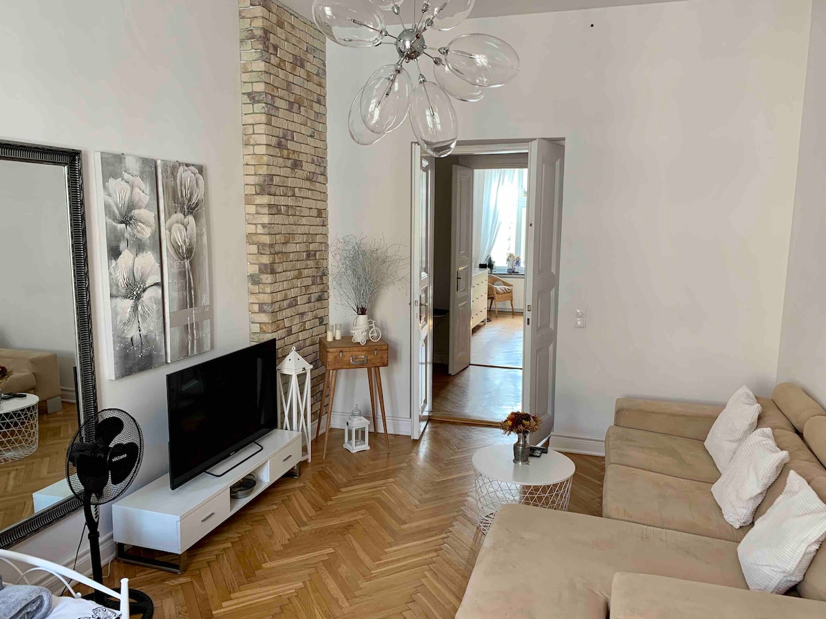 A living area features a light-colored sofa arranged around a small coffee table. A modern television is placed on a low cabinet. A decorative mirror and wall art add character, while wooden flooring and a brick accent wall create a cozy setting. Natural light fills the space through an open doorway.