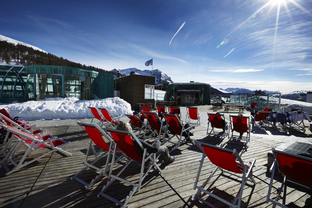 A spacious outdoor terrace is seen, featuring multiple red lounge chairs arranged on a wooden deck. Snow-covered mountains are visible in the background, along with a clear blue sky dotted with wispy clouds. Glimmers of sunlight reflect off the surrounding scenery.