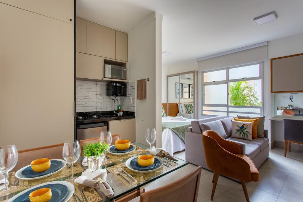 A modern kitchen area is visible, featuring sleek cabinetry and a compact dining table set for four. A comfortable sofa is positioned nearby, with a bed area and natural light filtering through large windows, showcasing greenery outside and enhancing the overall spaciousness.
