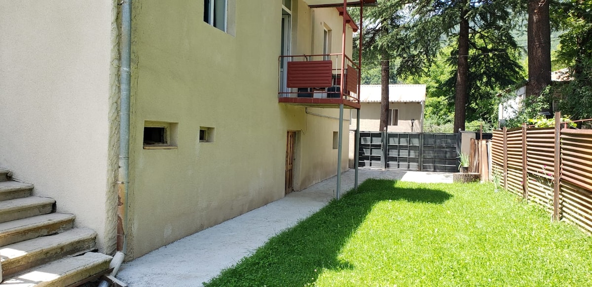 A view of the exterior of the building showcases a balcony adorned with a red railing. A well-maintained grassy area is flanked by smooth concrete pathways that lead to a fenced space, surrounded by tall trees in the background.