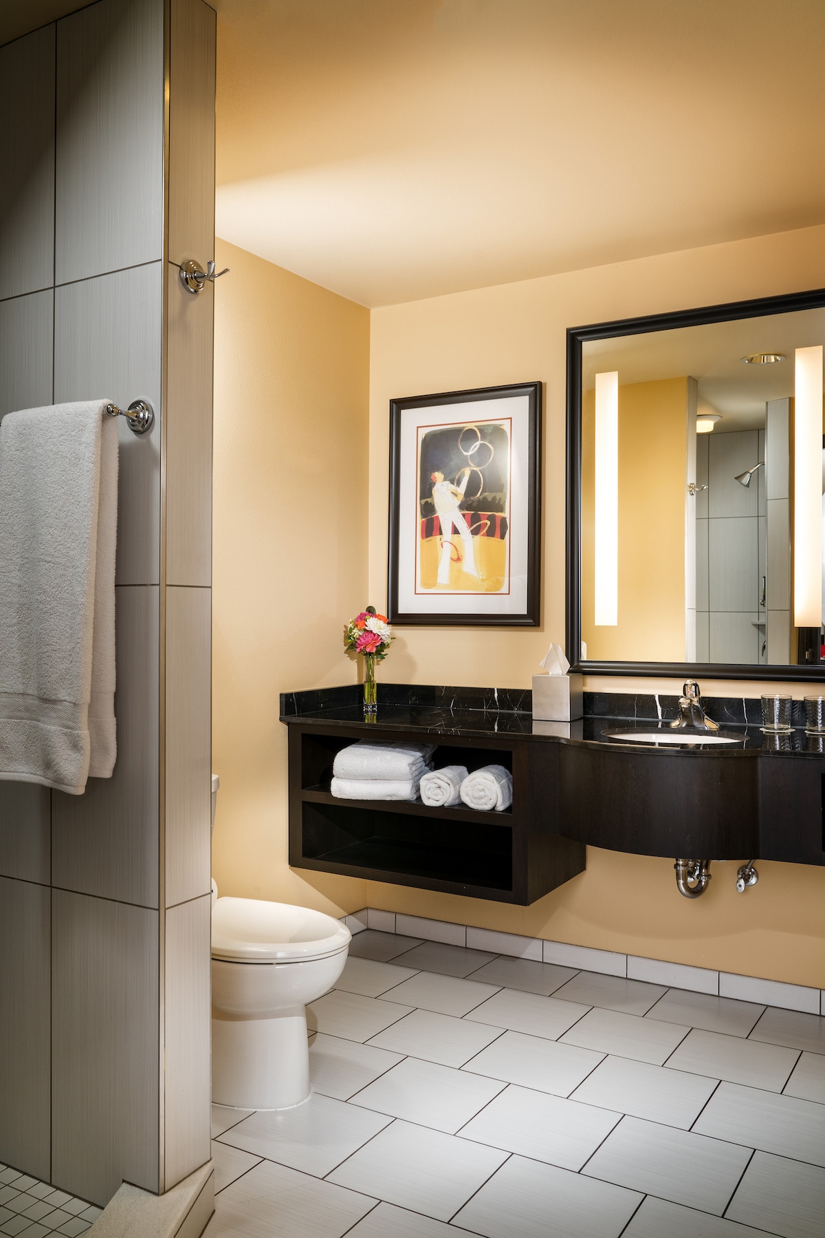 A bright bathroom features a walk-in shower with a glass partition. A large mirror is mounted above a dark vanity with neatly arranged towels. A flower arrangement adds a touch of color, while light beige walls complement the white tiled flooring.