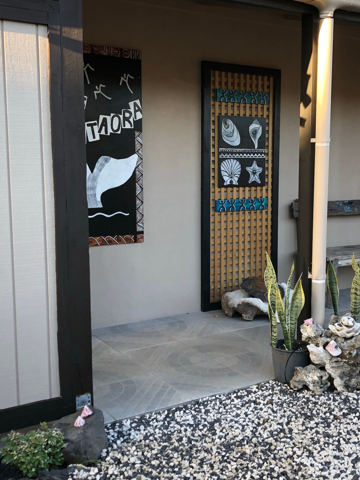 The entrance area features a decorative wall panel adorned with intricate carvings of marine life. A tiled pathway is bordered by decorative stones and plants, leading toward an inviting door that reflects the local culture.