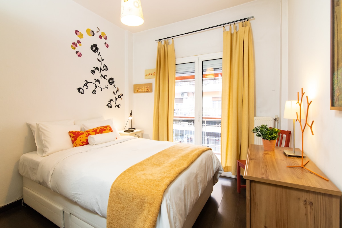 The bedroom features a spacious bed topped with white linens and a vibrant decorative throw. A decorative wall design of flowers adds a pop of color, and natural light filters through curtains. A small wooden desk and lamp provide a cozy workspace near the window.